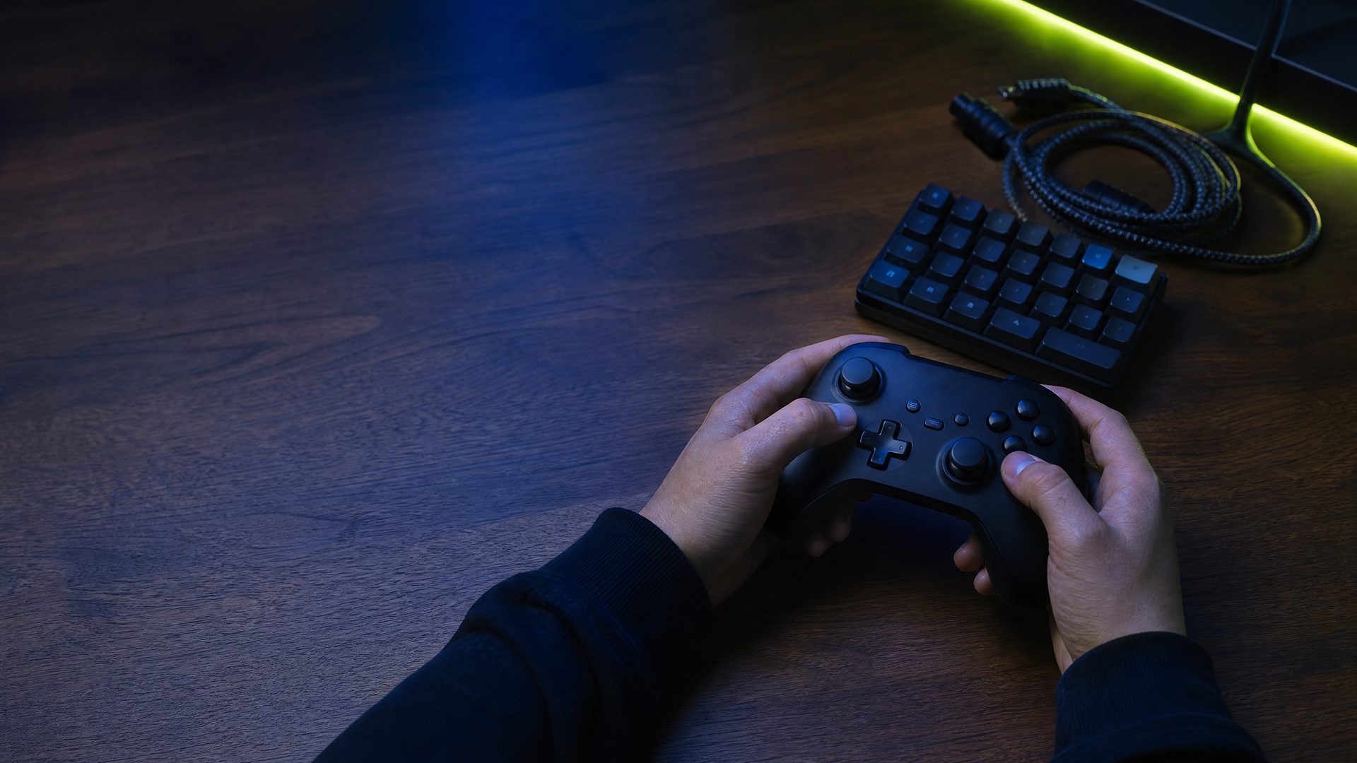 Two hands resting on a black game controller on a dark walnut desk under indigo light